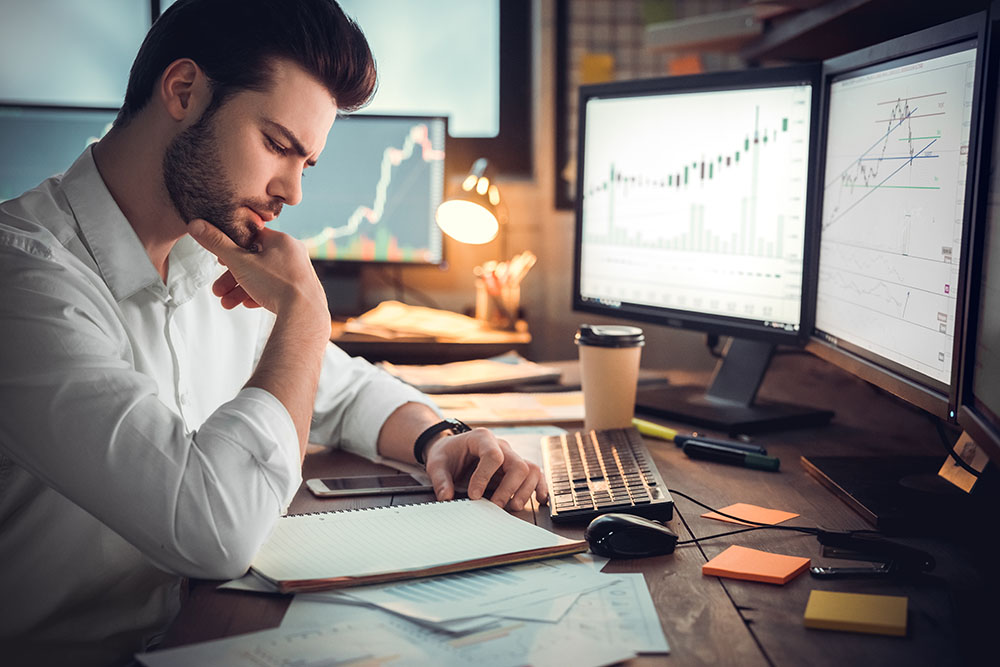 A guy wearing a white polo reading his trading journal and thinking which instruments he should trade. In front of him are 2 PC monitors with the graph or trading instruments.